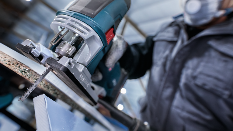 Person wearing safety equipment uses a jigsaw to cut a sheet of material.