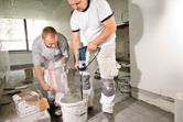 Two workers prepare mortar, one pours powder while the other mixes with a power drill.