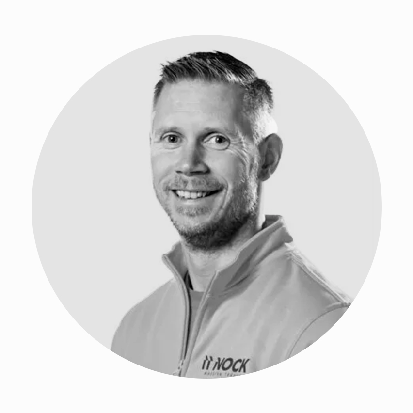 En leende man med kort hår i en grå jacka med logotyp.