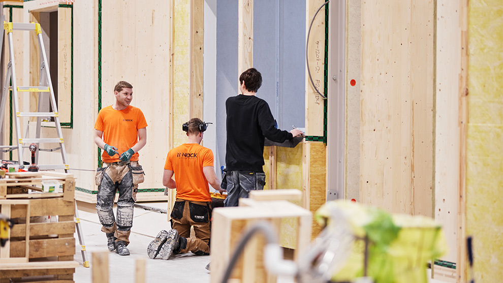 Tre hantverkare i orange tröjor arbetar med väggar i en byggnad.