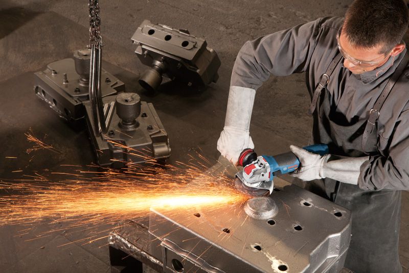 En person med säkerhetsutrustning slipar ett metallblock med en vinkelslip, vilket producerar gnistor.