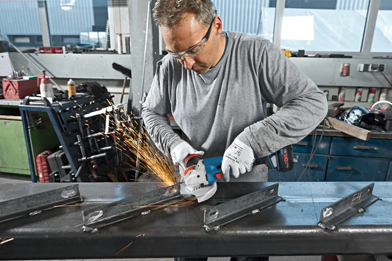 En arbetare med säkerhetsutrustning slipar metallbalkar med en sladdlös vinkelslip.