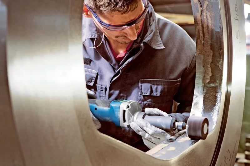 En person med säkerhetsutrustning polerar en stor metallring med en sladdlös rak slipmaskin.