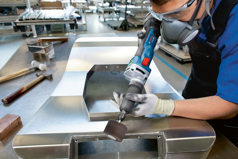 En person med säkerhetsutrustning slipar en metallplåt med en sladdlös rak slipmaskin.
