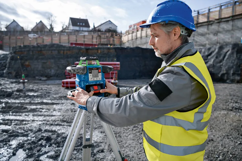 En person med säkerhetsutrustning justerar ett lasernivelleringsverktyg på ett stativ på en byggarbetsplats.