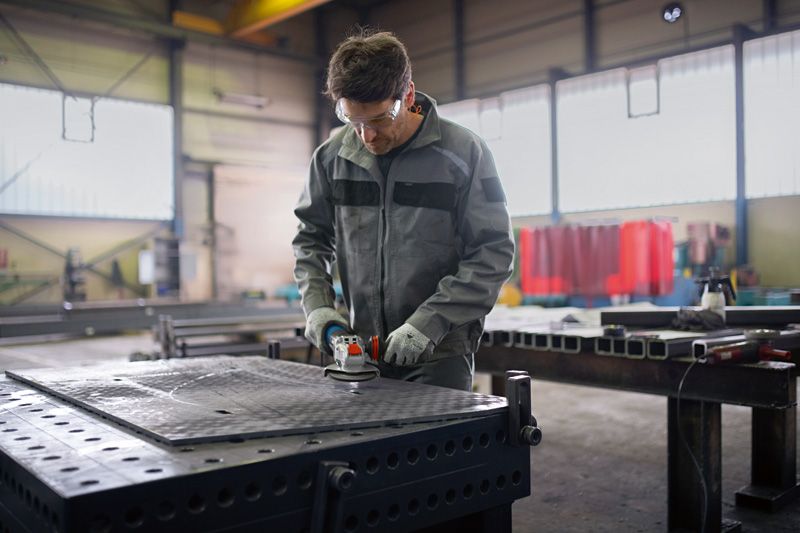 En person med säkerhetsutrustning använder en vinkelslip på ett arbetsbord av metall.