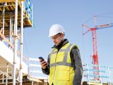 En person med säkerhetsutrustning kontrollerar en smartphone på en byggarbetsplats.