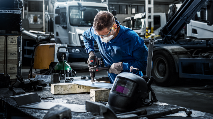 Arbetare med säkerhetsutrustning borrar metall på en arbetsbänk i ett industrigarage.