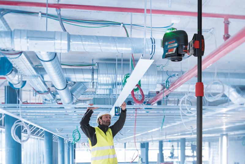 En person med säkerhetsutrustning installerar en takpanel med hjälp av ett lasernivelleringsverktyg på en teleskopstång.