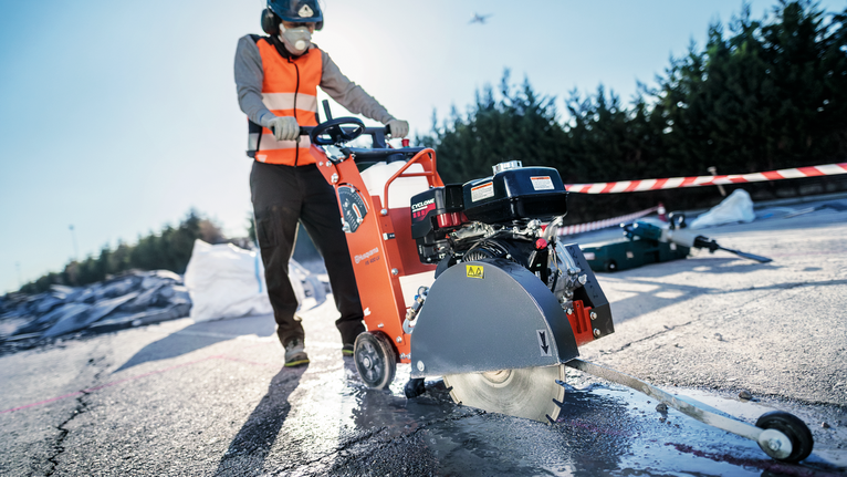 En person med säkerhetsutrustning använder en betongsåg på en asfalterad yta.