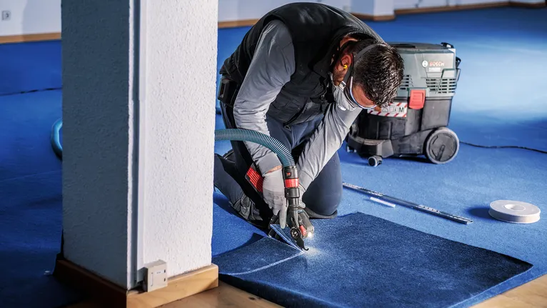 En person med säkerhetsutrustning skär i blå mattor med ett elverktyg inomhus.