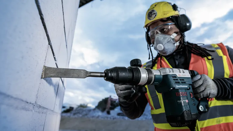 En person med säkerhetsutrustning mejslar en betongvägg med ett elverktyg.
