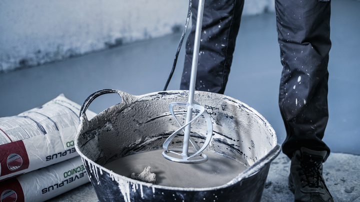 Person som bär säkerhetsutrustning blandar cement med en kraftblandare i en stor hink.