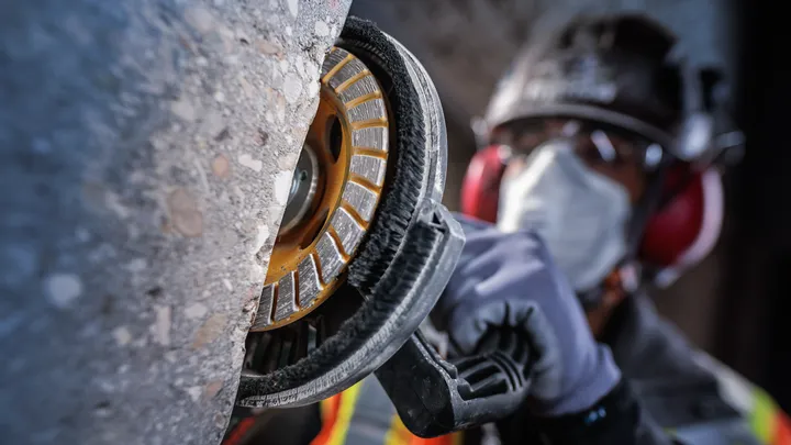 En person med säkerhetsutrustning slipar en betongvägg med ett elverktyg.