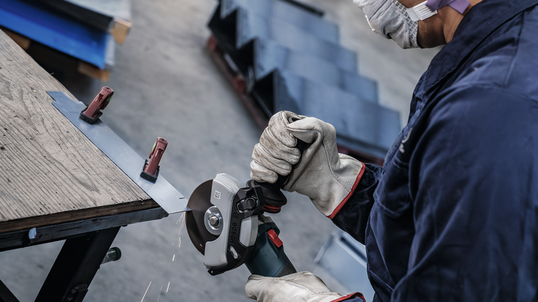 En person med säkerhetsutrustning slipar en metallplåt som är fastklämd på en arbetsbänk.