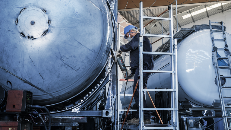 En person med säkerhetsutrustning använder ett elverktyg på en stor metalltank.
