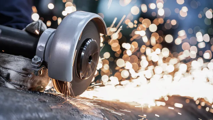 En person som bär säkerhetsutrustning använder en vinkelslip för att skära metall, vilket skapar gnistor.