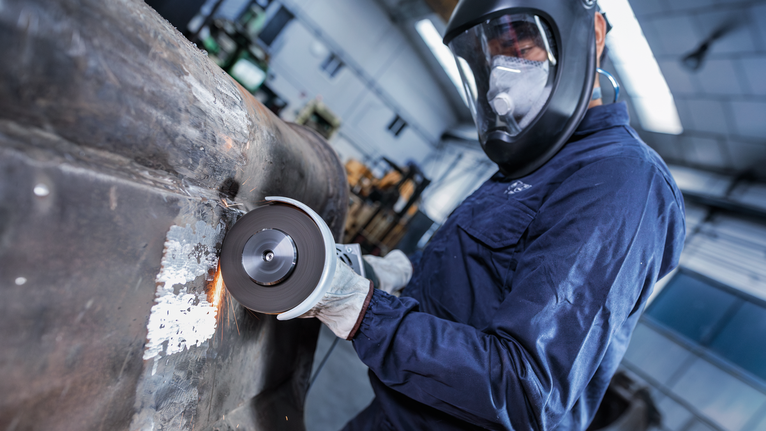 En person som bär säkerhetsutrustning slipar en stor metallyta i en industriell verkstad.