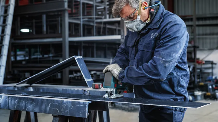 En person som bär säkerhetsutrustning använder ett elverktyg för att skära en metallplåt.