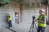 Två personer med säkerhetsutrustning ställer upp ett lasernivelleringsverktyg på ett stativ i ett betongrum.