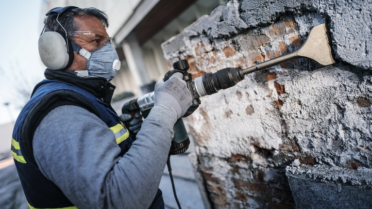 Person med säkerhetsutrustning mejslar betong från en tegelvägg med ett elverktyg.