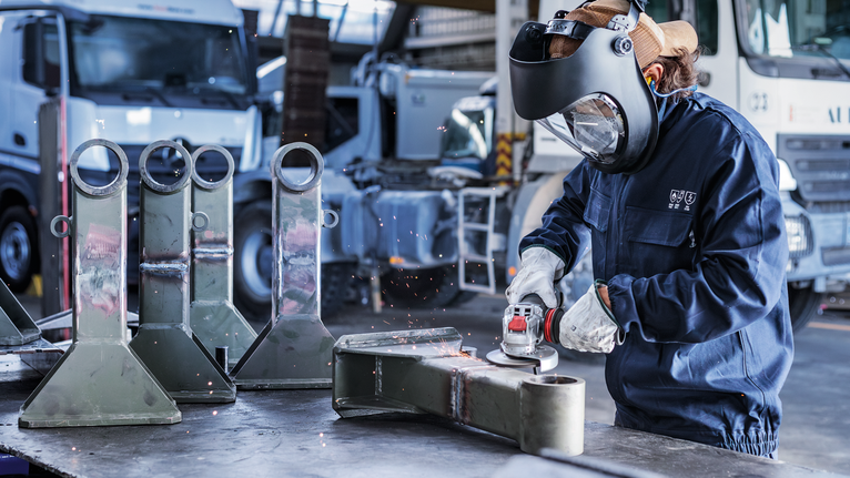 En person som bär säkerhetsutrustning slipar en metalldel i en industriell miljö.