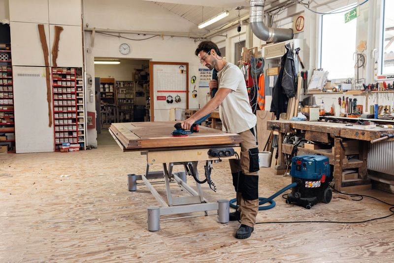 En person med säkerhetsutrustning slipar en träyta med en sladdlös excenterslip.