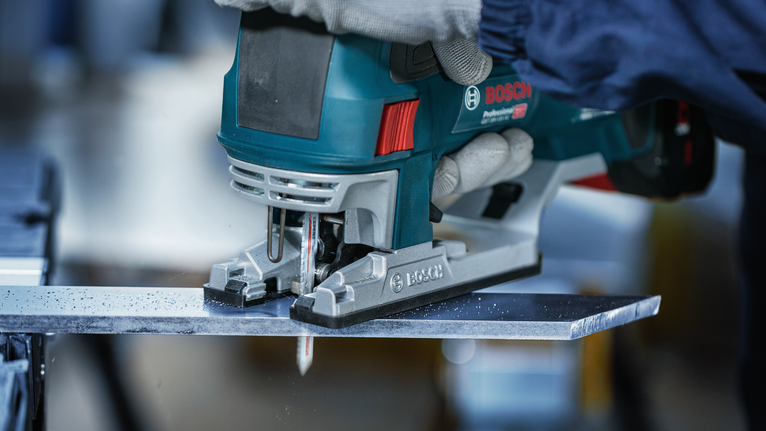 En person som bär säkerhetsutrustning sågar en metallplåt med en handhållen sticksåg.