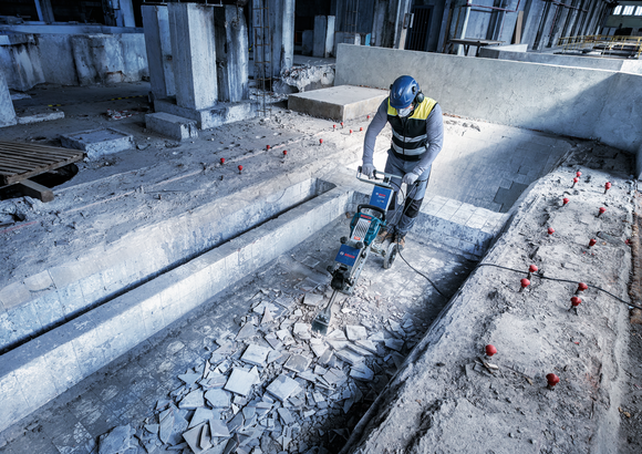 En person med säkerhetsutrustning använder en rivningshammare för att slå sönder betongplattor.