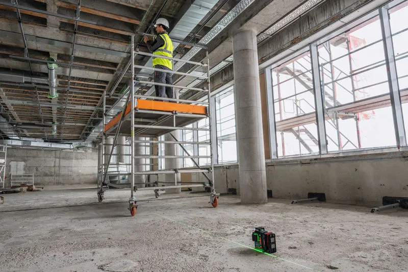 En person som bär säkerhetsutrustning arbetar på en byggnadsställning med ett lasernivelleringsverktyg på golvet.