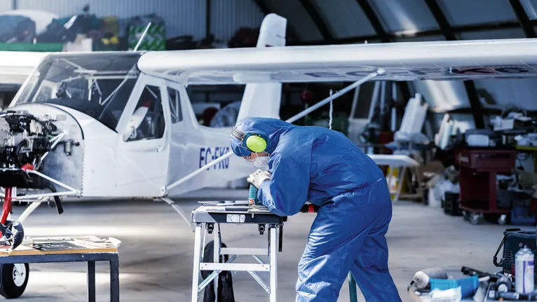 En person som bär säkerhetsutrustning borrar en metalldel på en arbetsbänk nära ett litet flygplan.