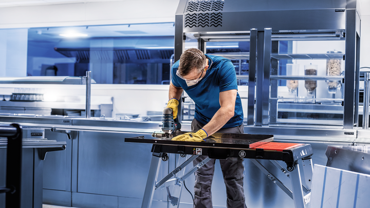 En person med säkerhetsutrustning slipar en metallplåt på en arbetsbänk i ett kök.