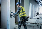 En person med säkerhetsutrustning borrar i gipsväggar nära ett lasernivelleringsverktyg på ett stativ.