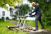 En person med säkerhetsutrustning använder en sladdlös motorsåg för att såga trädgrenar utomhus.