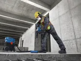 En arbetare med säkerhetsutrustning använder en borrhammare på ett betonggolv.