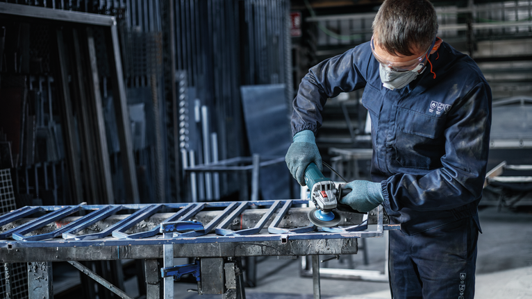 En person med säkerhetsutrustning slipar metallstänger i en industriell verkstad.