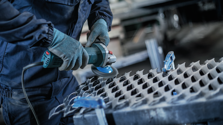 En person som bär säkerhetsutrustning använder en vinkelslip på en metallplåt.