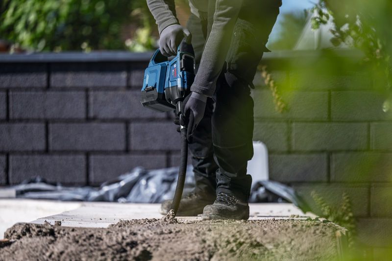 En person med säkerhetsutrustning använder ett elverktyg för att borra i betong utomhus.