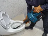 En person som bär säkerhetsutrustning använder ett elverktyg för att rensa en toalett.