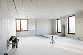 Person som knäböjer för att mäta en vägg medan lasernivelleringsverktyg projicerar linjer över ett ljust rum.
