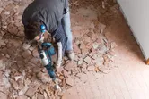 Oseba v varnostni opremi lomi talne ploščice z rušilnim kladivom.
