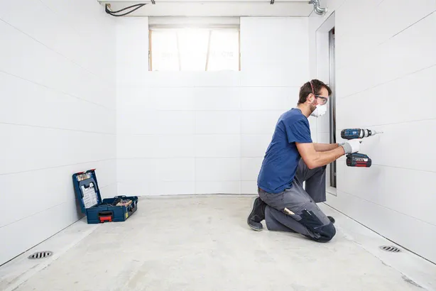 Oseba, ki nosi varnostno opremo, vrta v ploščice obloženo steno v prazni sobi.