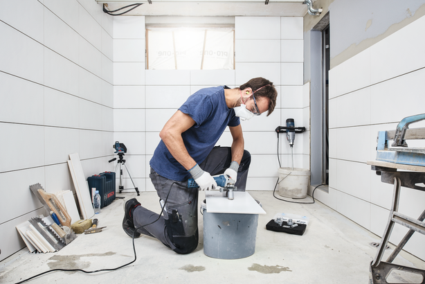 Oseba v varnostni opremi izvrta luknjo v ploščico v prostoru s ploščicami.