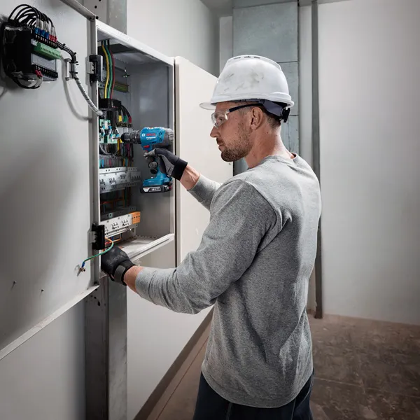 Oseba, ki nosi varnostno opremo, uporablja akumulatorski vrtalnik na električni plošči.