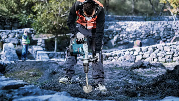 Oseba, ki nosi varnostno opremo, uporablja vrtalno kladivo za kopanje tal na prostem.