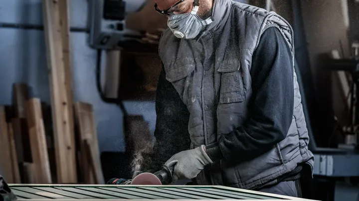 Oseba, ki nosi varnostno opremo, brusi leseno površino z električnim orodjem.