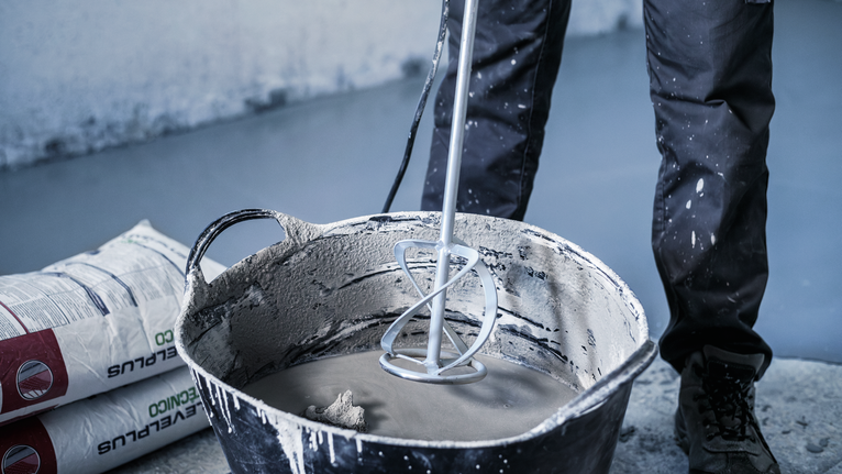 Oseba v varnostni opremi meša cement z električnim mešalnikom v velikem vedru.