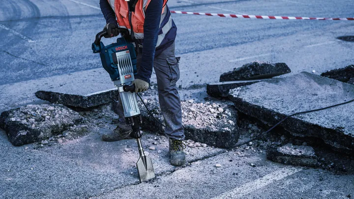 Oseba v varnostni opremi lomi asfalt z rušilnim kladivom.