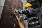 Oseba, ki nosi varnostno opremo, brusi zarjavelo kovinsko pločevino z akumulatorskim kotnim brusilnikom.