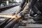 Oseba v varnostni opremi reže železniške tire z akumulatorskim kotnim brusilnikom.
